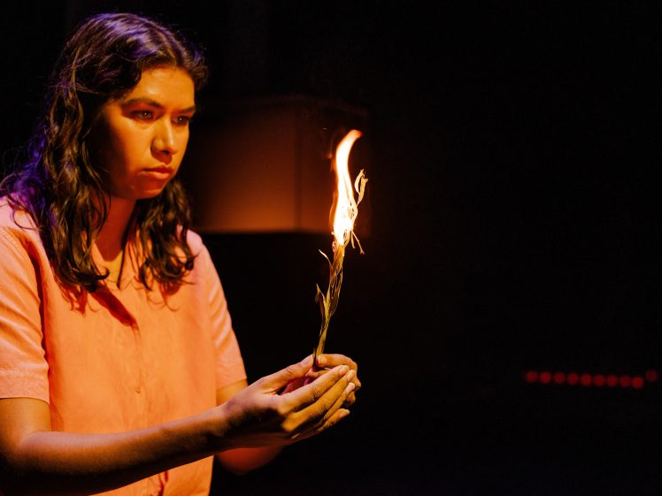 Image of A person with long, wavy hair and a serious expression holds a small burning branch or stick, with bright flames rising, against a dark background.