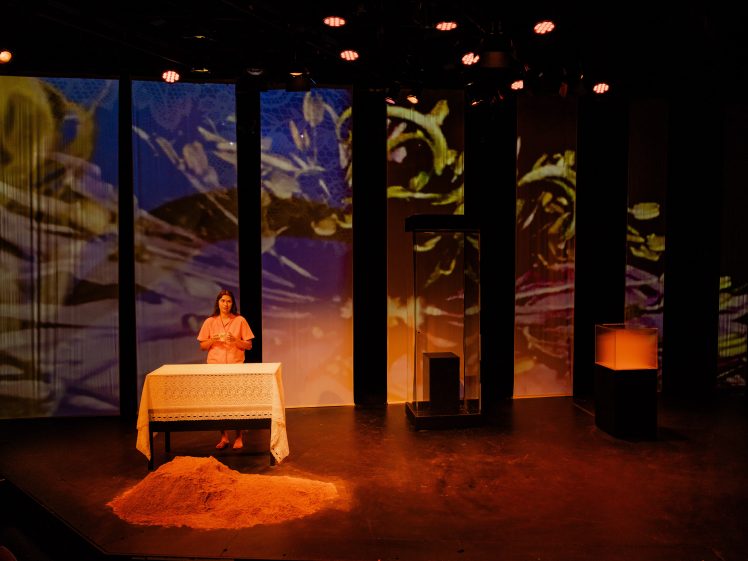 Image of A woman stands behind a table covered with a white cloth on a stage. There is a pile of sand in front of the table, and abstract, colourful visuals are projected on panels behind her. The lighting is dramatic and warm.