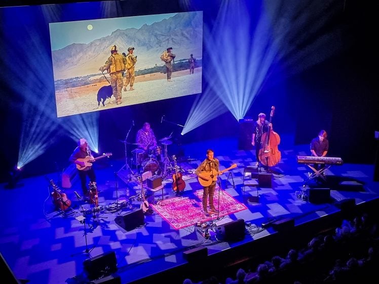 Image of A band performs on a stage with guitars, drums, keyboard, and a double bass. Blue stage lights shine, and a large screen behind shows soldiers walking in a desert landscape with mountains.