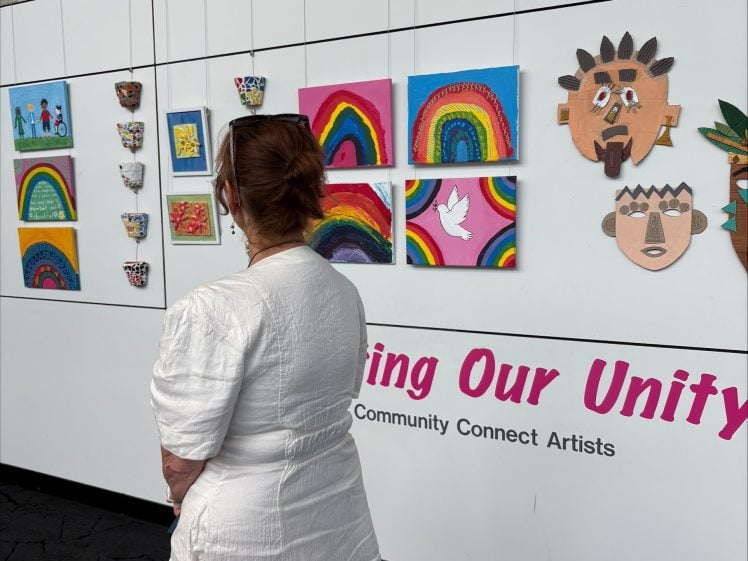 Image of A woman in a white dress stands in front of a display of colourful children’s artwork, including rainbows, doves, masks, and small decorated pots, mounted on a white wall.